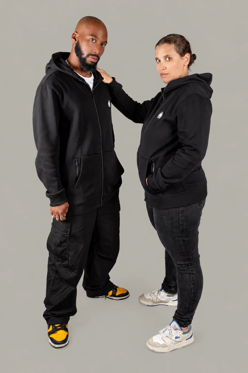 Deux personnes portant le hoodie Origins noir, sur fond gris taupe neutre